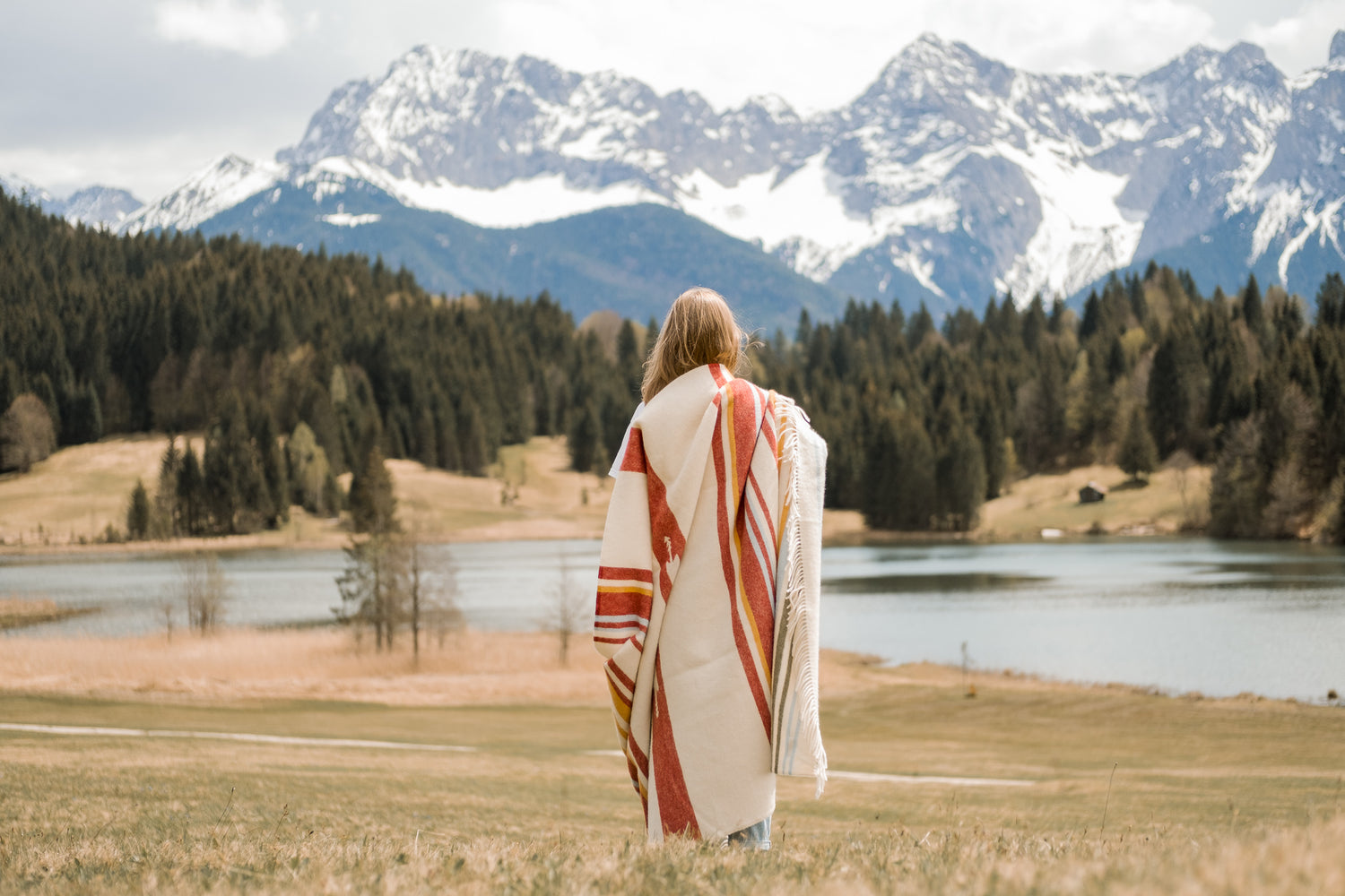 Eine junge Frau steht mit einer bunten Tribal decke auf einer Alm mit Bergpanorama