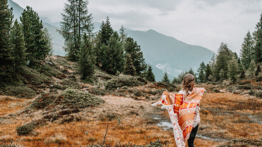 Eine bunte Schurwolldecke mit zickzack Muster und Fransen liegt auf einer Holzbank in der Natur.