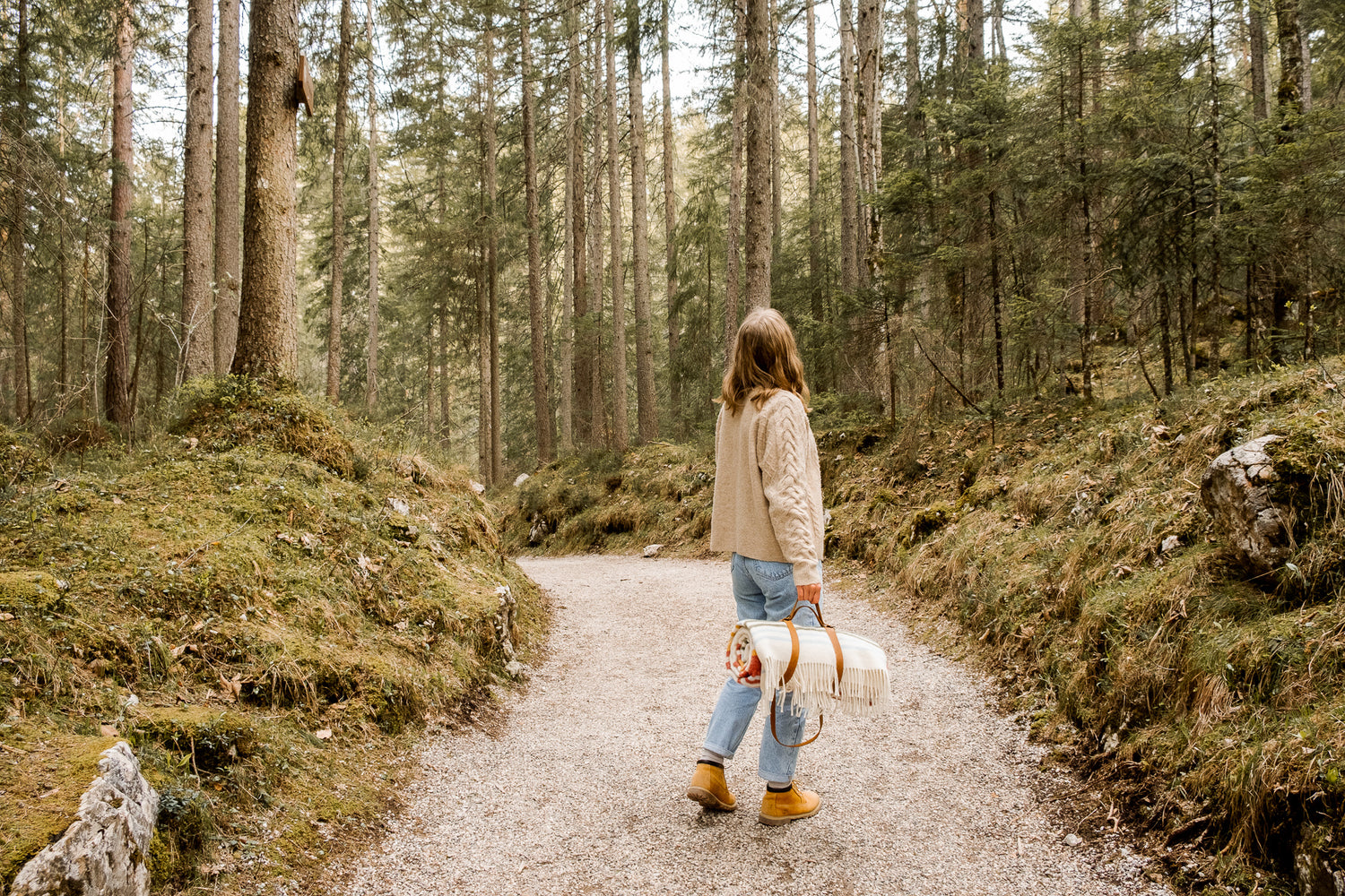 Eine junge Wanderin läuft durch den Wald und hält in einer Hand eine bunte Wolldecke mit Fransen und Lederriemengeschirr 