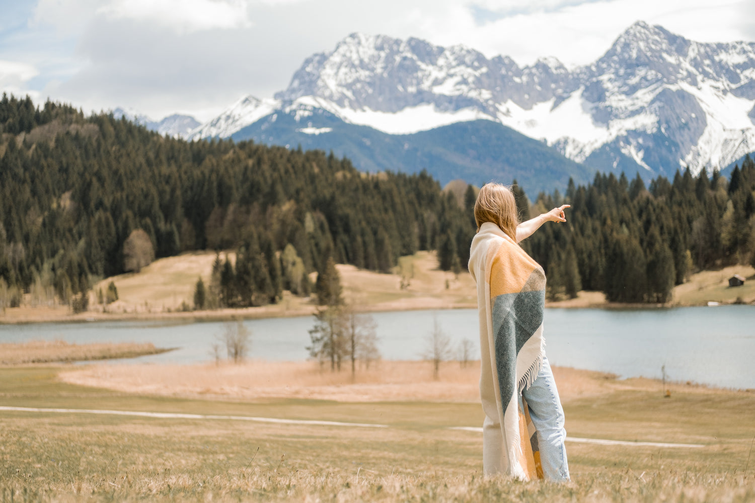 Eine junge Frau trägt eine weiche Wolldecke aus ökologischer Schafwolle auf dem Arm und zeigt auf ein schönes Bergpanorama in den Alpen  